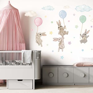 K&ouml;nnte beinhalten: Ein wei&szlig;es Babybett mit einem rosa Baldachin und eine graue Kommode mit einer wei&szlig;en Schublade. Die Wand hinter dem Babybett ist mit einem Wandtattoo f&uuml;r Kinderzimmer dekoriert, das braune Hasen zeigt, die pastellfarbene Ballons und Wolken halten.