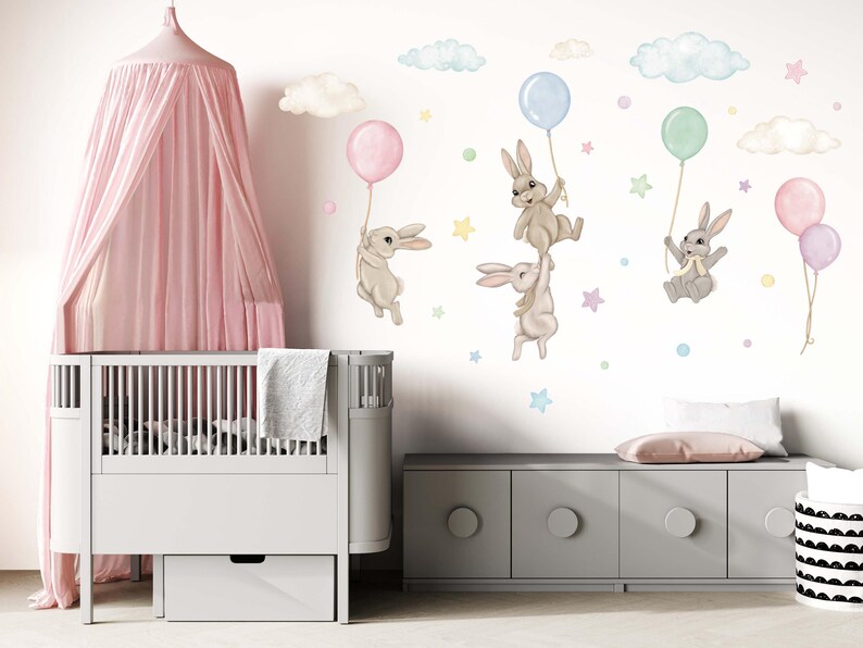 K&ouml;nnte beinhalten: Ein wei&szlig;es Babybett mit einem rosa Baldachin und eine graue Kommode mit einer wei&szlig;en Schublade. Die Wand hinter dem Babybett ist mit einem Wandtattoo f&uuml;r Kinderzimmer dekoriert, das braune Hasen zeigt, die pastellfarbene Ballons und Wolken halten.