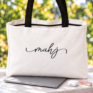 May include: A white canvas tote bag with black fabric handles and a black script graphic that reads mahj with a small red heart above the letter j. The bag features a rectangular shape and a clean, simple design for carrying items.