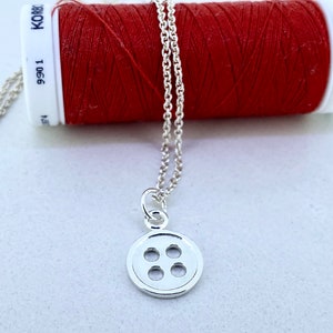 May include: A silver necklace with a button charm. The button has four holes and is round. The necklace is on a white surface next to a spool of red thread.