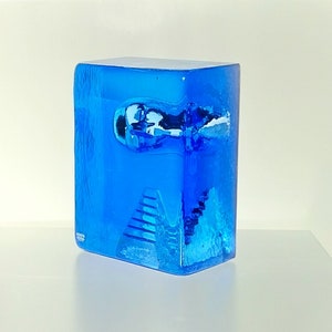 May include: A striking blue art glass sculpture, rectangular in form. The glass has a textured surface with an abstract design and a small, rounded indentation. A staircase design is visible at the base of the sculpture.