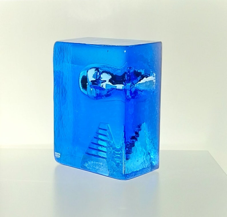 May include: A striking blue art glass sculpture, rectangular in form. The glass has a textured surface with an abstract design and a small, rounded indentation. A staircase design is visible at the base of the sculpture.