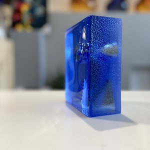 May include: A rectangular, transparent block of vibrant blue glass. The glass has a textured surface and appears to have embedded objects within. The block is resting on a white surface, with a blurred background.