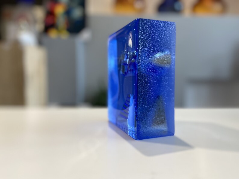 May include: A rectangular, transparent block of vibrant blue glass. The glass has a textured surface and appears to have embedded objects within. The block is resting on a white surface, with a blurred background.