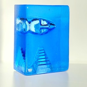 May include: A striking blue glass sculpture featuring a fish and pyramid design. This Kosta Boda art glass piece has a rectangular form with a textured surface and a signature. The fish is white and blue.