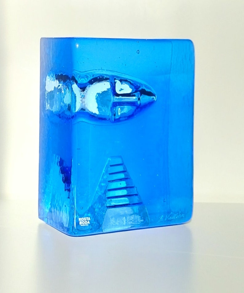 May include: A striking blue glass sculpture featuring a fish and pyramid design. This Kosta Boda art glass piece has a rectangular form with a textured surface and a signature. The fish is white and blue.