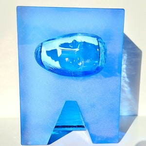May include: A blue glass sculpture with a rectangular base and a rounded, oval-shaped element in the centre. The sculpture has a triangular cutout at the bottom. The glass has a frosted appearance.