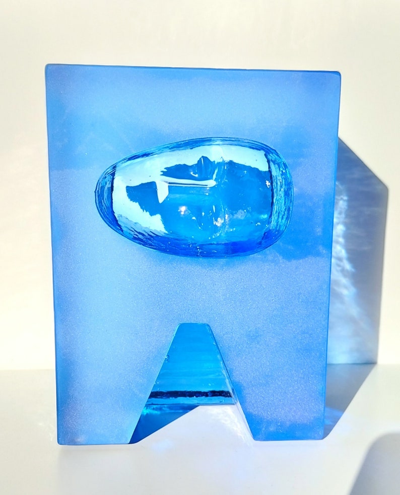 May include: A blue glass sculpture with a rectangular base and a rounded, oval-shaped element in the centre. The sculpture has a triangular cutout at the bottom. The glass has a frosted appearance.
