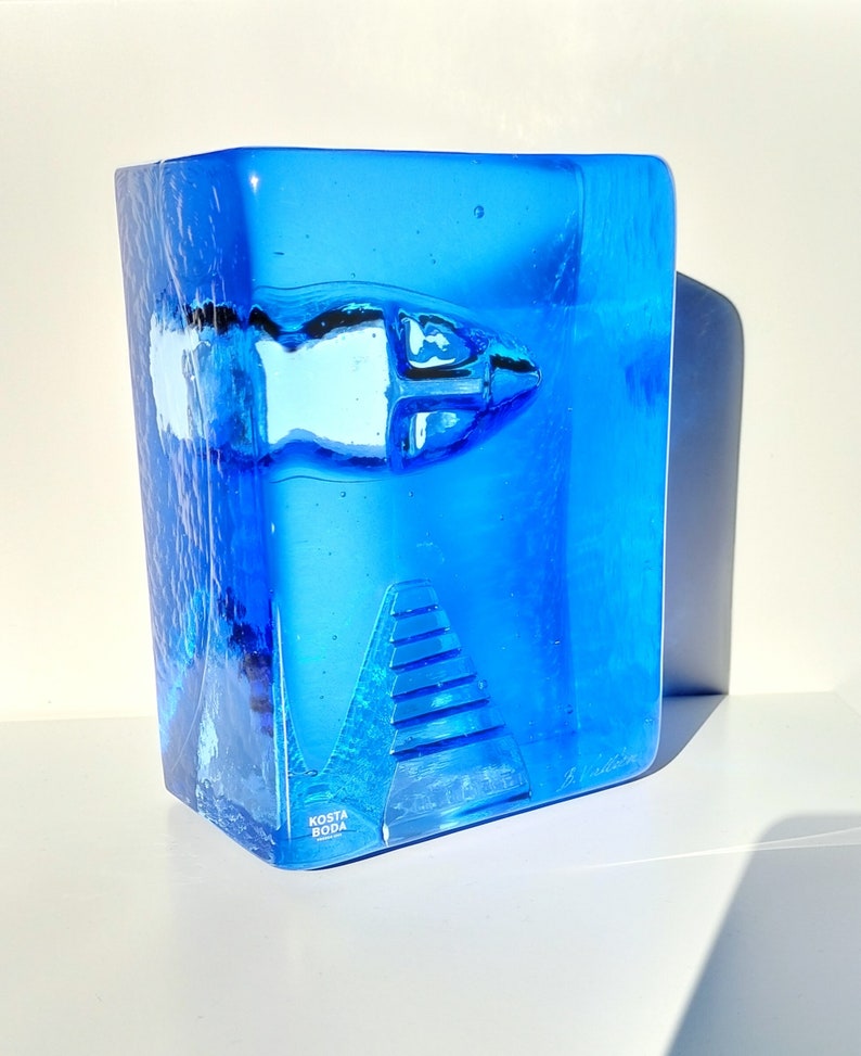 May include: A striking blue glass sculpture featuring a fish design and a textured finish. The rectangular sculpture has a fish motif and a pyramid-like structure. The base of the sculpture has the text "KOSTA BODA".