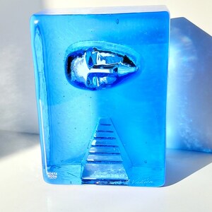 May include: A striking blue glass sculpture by Kosta Boda. The rectangular form has an embedded oval design and a triangular, stepped feature. The sculpture is signed and has a clear, transparent quality, with light and shadow effects.