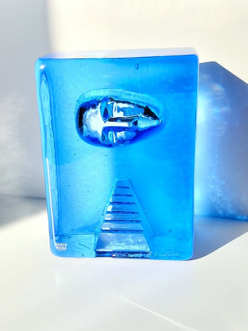 May include: A striking blue glass sculpture by Kosta Boda. The rectangular form has an embedded oval design and a triangular, stepped feature. The sculpture is signed and has a clear, transparent quality, with light and shadow effects.