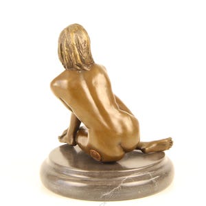 May include: Bronze sculpture of a seated figure, viewed from the back. The figure is positioned on a dark, circular base. The sculpture has a smooth, polished finish, with details in the hair and form.