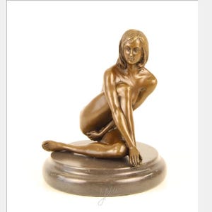 May include: Bronze sculpture of a seated woman with long hair, resting on a circular base. The figure is posed with one leg extended and arms gracefully positioned. The sculpture has a warm, metallic brown color.