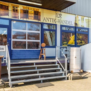 May include: Exterior shot of an antique shop with a sign that reads "Antiquit&auml;ten". The shop has blue trim and large windows. A small wooden stool holds a round object. The shop's name, "ANTIK-HANDEL", is displayed above the windows.