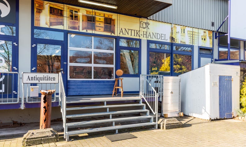May include: Exterior shot of an antique shop with a sign that reads "Antiquit&auml;ten". The shop has blue trim and large windows. A small wooden stool holds a round object. The shop's name, "ANTIK-HANDEL", is displayed above the windows.