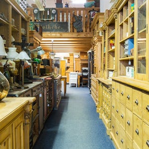 May include: Interior shot of a vintage shop with antique furniture and decor. The shop features wooden cabinets, drawers, and shelves filled with various items. A large glass bottle with a cork stopper sits on a wooden counter.