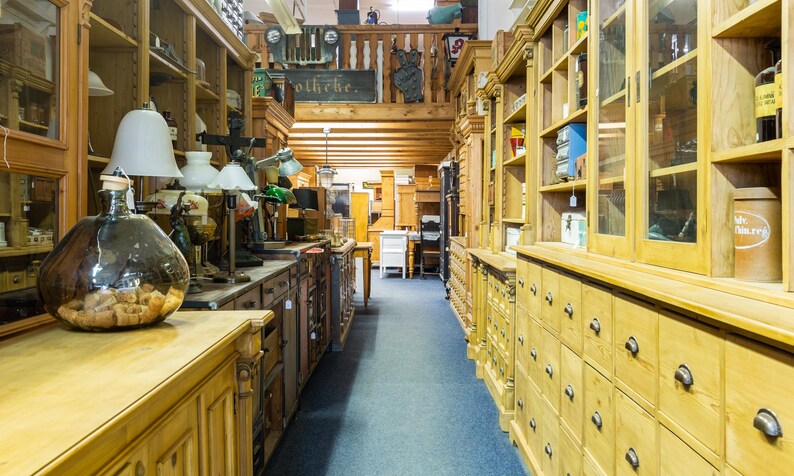 May include: Interior shot of a vintage shop with antique furniture and decor. The shop features wooden cabinets, drawers, and shelves filled with various items. A large glass bottle with a cork stopper sits on a wooden counter.
