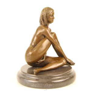 May include: A bronze sculpture of a seated figure with short hair, resting on a round, dark gray base. The figure is nude, with arms and legs crossed. The sculpture has a smooth, polished finish and is set against a white background.