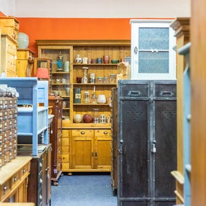 May include: A shop interior filled with antique furniture. Wooden cabinets, shelves, and dressers in various styles and colors are visible. A blue painted shelf and a pair of dark metal storage cabinets are also present. The walls are painted orange.
