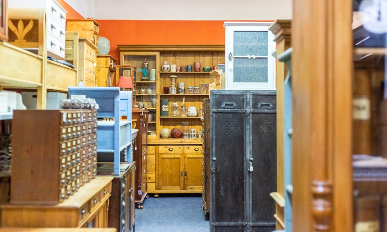 May include: A shop interior filled with antique furniture. Wooden cabinets, shelves, and dressers in various styles and colors are visible. A blue painted shelf and a pair of dark metal storage cabinets are also present. The walls are painted orange.