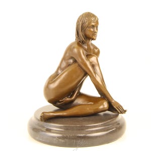 May include: Bronze sculpture of a seated figure with a serene expression. The figure is posed with one leg bent and the other extended, resting on a circular base. The sculpture has a smooth, polished finish and is set against a white background.