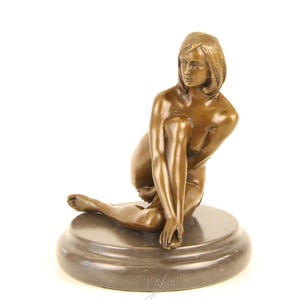 May include: A bronze sculpture of a seated figure with short hair, resting on a circular base. The figure is posed with one leg extended and the other bent, with the arms gently touching the legs. The sculpture has a smooth, polished finish.