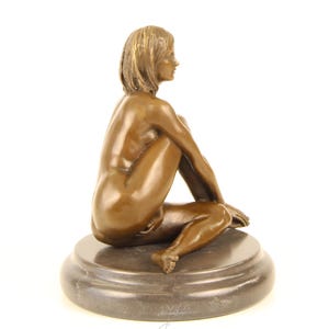 May include: Bronze sculpture of a seated figure with short hair, resting on a round, dark base. The figure is nude, with arms and legs crossed. The sculpture has a smooth, polished finish and is set against a white background.