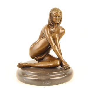 May include: Bronze sculpture of a seated figure with crossed arms and legs. The figure is positioned on a dark, circular base. The sculpture has a smooth, polished finish and is set against a white background.