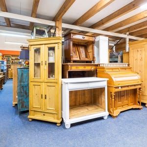 May include: A showroom filled with various wooden furniture pieces. The collection includes cabinets, desks, and wardrobes in different styles and finishes. The floor is covered in blue carpet, and the ceiling has exposed wooden beams.
