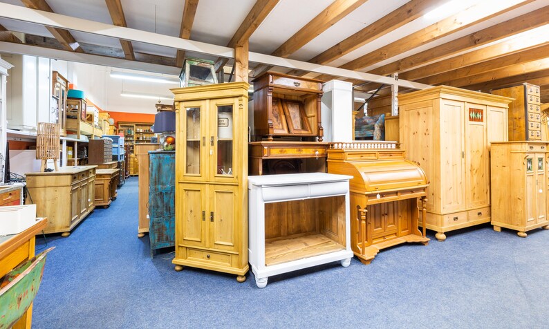 May include: A showroom filled with various wooden furniture pieces. The collection includes cabinets, desks, and wardrobes in different styles and finishes. The floor is covered in blue carpet, and the ceiling has exposed wooden beams.