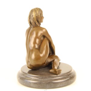May include: Bronze sculpture of a seated figure with short hair, positioned with their back to the viewer. The figure is on a round, dark gray marble base. The sculpture has a smooth, polished finish and is set against a white background.