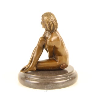 May include: Bronze sculpture of a seated figure with short hair, resting on a dark, circular base. The figure is in a relaxed pose, with one leg bent and the other extended. The sculpture has a smooth, polished finish.