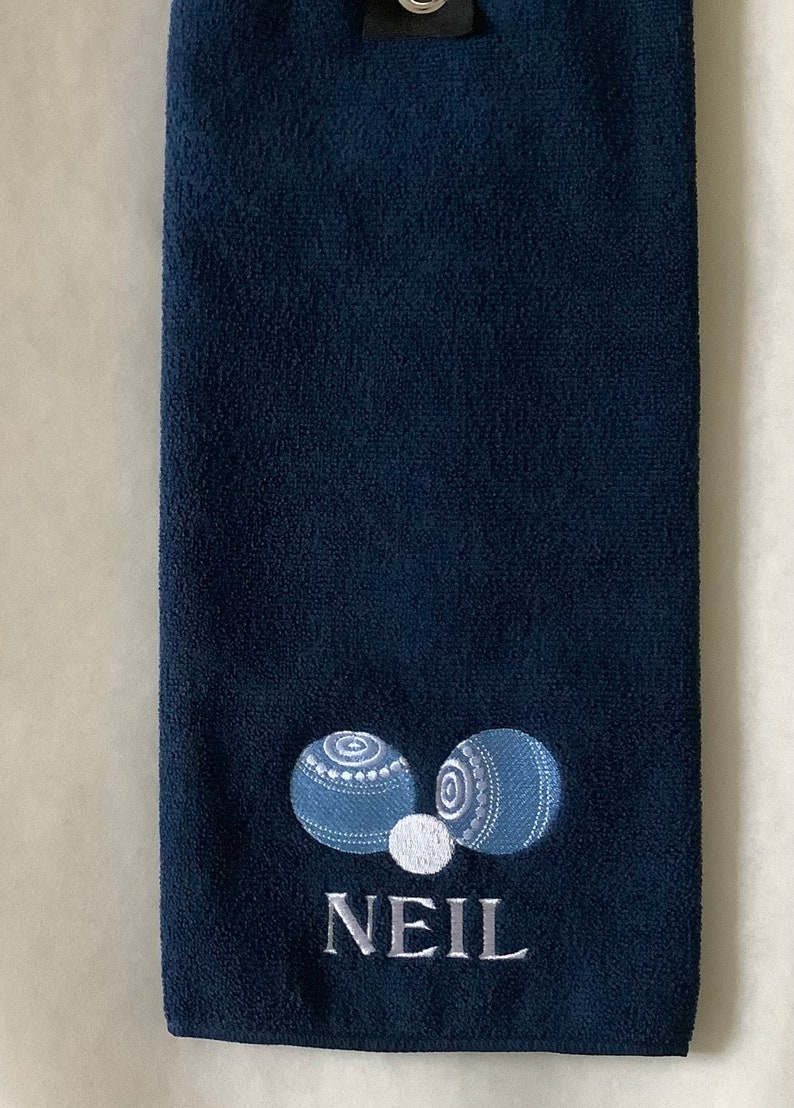 K&ouml;nnte beinhalten: Ein dunkelblaues Handtuch mit einem wei&szlig;en gestickten Design aus zwei blauen Bowlingkugeln und einer wei&szlig;en Bowlingkugel. Der Name "NEIL" ist unter den Bowlingkugeln gestickt.