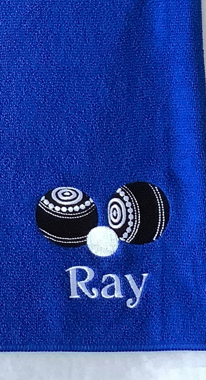 K&ouml;nnte beinhalten: Ein blaues Handtuch mit zwei wei&szlig;en und schwarzen Bowlingkugeln und dem Namen "Ray" bestickt.