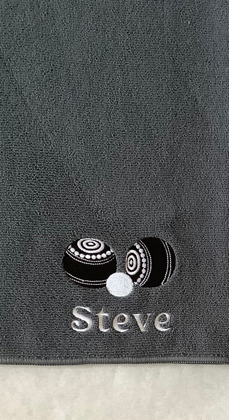 K&ouml;nnte beinhalten: Ein graues Handtuch mit zwei schwarzen und wei&szlig;en Bowlingkugeln und dem Namen "Steve", der in Wei&szlig; gestickt ist.