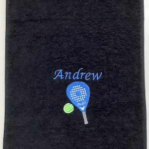 May include: A black rectangular towel featuring an embroidered design in the centre. The embroidery includes the name Andrew in blue script, a blue padel racket with white detailing, and a small lime green padel ball positioned to the left.