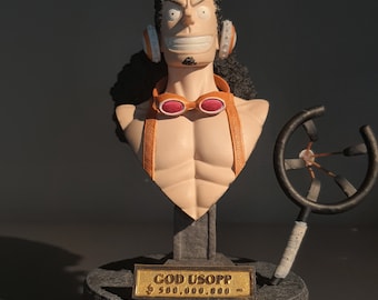 May include: A bust of a cartoon character with brown hair and a goatee, wearing goggles and a brown strap across his chest. The bust is on a black base with a gold plaque that reads "GOD USOPP $ 500,000,000" and a black and white sign that reads "DEAD OR ALIVE WANTED MARINE".