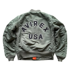 May include: An olive green bomber jacket featuring a ribbed collar, cuffs, and hem. The back displays the text AVIREX USA in large, dark navy blue embroidered letters with white outlines. The jacket has long sleeves and a visible orange zipper pull.