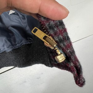 Puede incluir: Primer plano de una cremallera dorada de chaqueta con las letras "YKK" estampadas. La tela de la chaqueta es una mezcla de gris, negro y burdeos. El forro interior es azul oscuro.