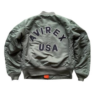 May include: An olive green bomber jacket featuring the text AVIREX USA in dark navy blue and white embroidered lettering on the back. The jacket has a ribbed collar, cuffs, and hem, with a small orange fabric detail visible at the bottom zipper.
