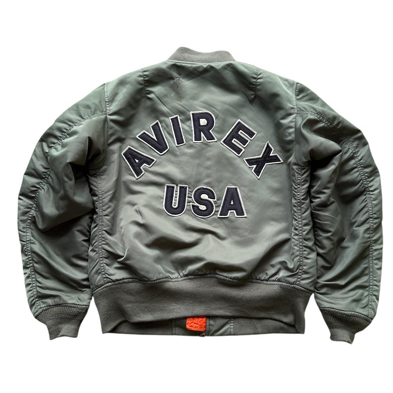 May include: An olive green bomber jacket featuring the text AVIREX USA in dark navy blue and white embroidered lettering on the back. The jacket has a ribbed collar, cuffs, and hem, with a small orange fabric detail visible at the bottom zipper.