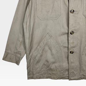 K&ouml;nnte beinhalten: Beige Jacke mit Knopfverschluss, zwei Brusttaschen und einer gro&szlig;en unteren Tasche. Die Jacke hat eine lockere Passform und einen l&auml;ssigen Stil. Der Stoff scheint eine leichte Baumwoll- oder Leinenmischung zu sein.