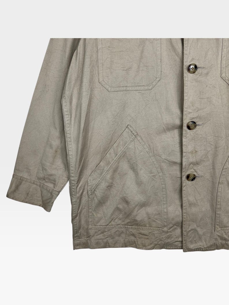 K&ouml;nnte beinhalten: Beige Jacke mit Knopfverschluss, zwei Brusttaschen und einer gro&szlig;en unteren Tasche. Die Jacke hat eine lockere Passform und einen l&auml;ssigen Stil. Der Stoff scheint eine leichte Baumwoll- oder Leinenmischung zu sein.