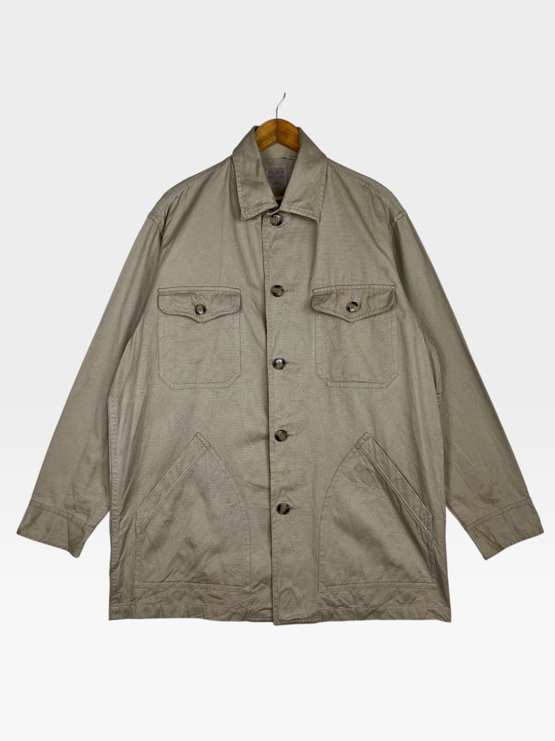 K&ouml;nnte beinhalten: Beigefarbene Jacke mit Knopfleiste, zwei Brusttaschen und zwei unteren Taschen. Die Jacke h&auml;ngt an einem Holzb&uuml;gel. Die Jacke hat einen Kragen und lange &Auml;rmel.