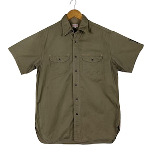 Può includere: Camicia verde oliva a maniche corte con due tasche sul petto e chiusura con bottoni. La camicia ha un colletto ed è appesa a una gruccia di legno. Il tessuto presenta un sottile motivo a righe verticali.