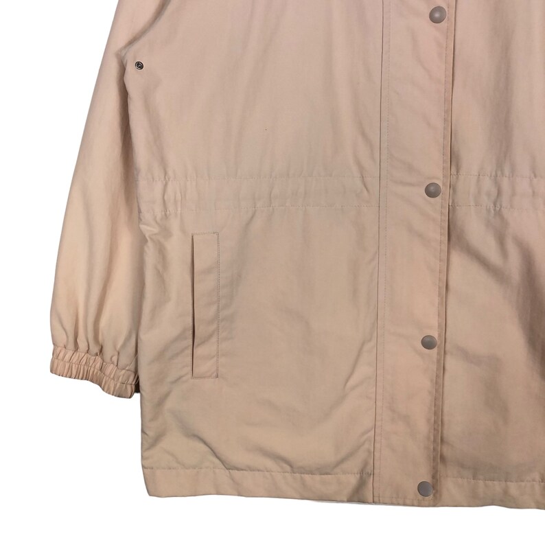 May include: A light beige jacket with a zipper and button closure. The jacket has a drawstring waist and a pocket on the left side.