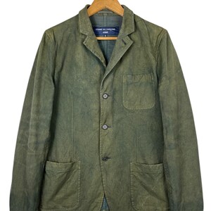 May include: Olive green blazer with three buttons and two front pockets. The jacket is hanging on a wooden hanger. The label on the collar reads "COMME des GAR&Ccedil;ONS HOMME".