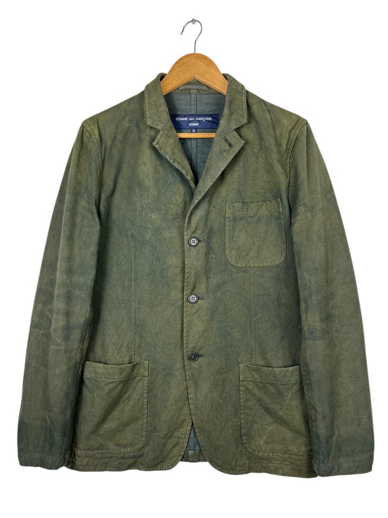May include: Olive green blazer with three buttons and two front pockets. The jacket is hanging on a wooden hanger. The label on the collar reads "COMME des GAR&Ccedil;ONS HOMME".