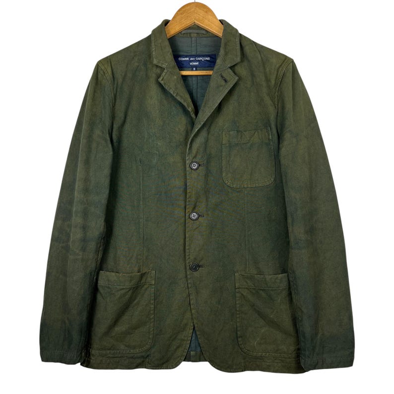May include: Olive green blazer with three buttons, two front pockets, and a chest pocket. The blazer is hanging on a wooden hanger. The label reads "COMME des GAR&Ccedil;ONS".