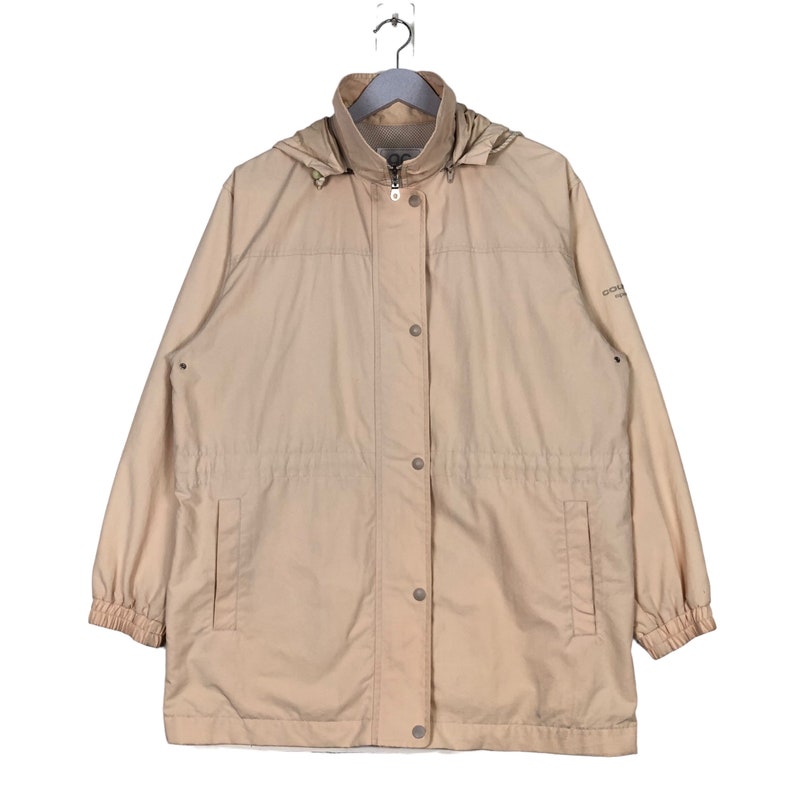 May include: A beige hooded jacket with a zipper and button closure. The jacket has a drawstring waist and two pockets. The jacket has the text "Columbia Sportswear Company" on the right sleeve.